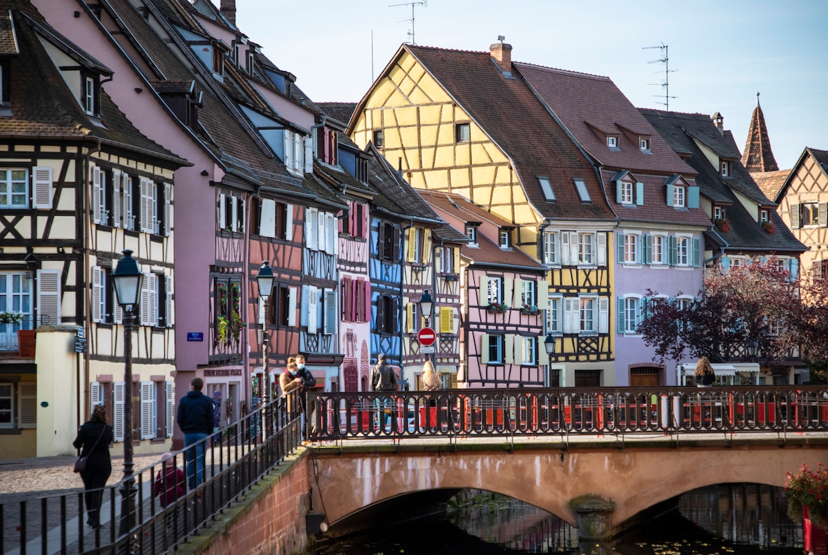 A charming street scene in Colmar features a row of half-timbered houses with pastel-colored facades lining the canal. A bridge crosses over the water, with pedestrians strolling leisurely. The area is characterized by its picturesque architecture and vibrant atmosphere, showcasing the unique Alsace region.