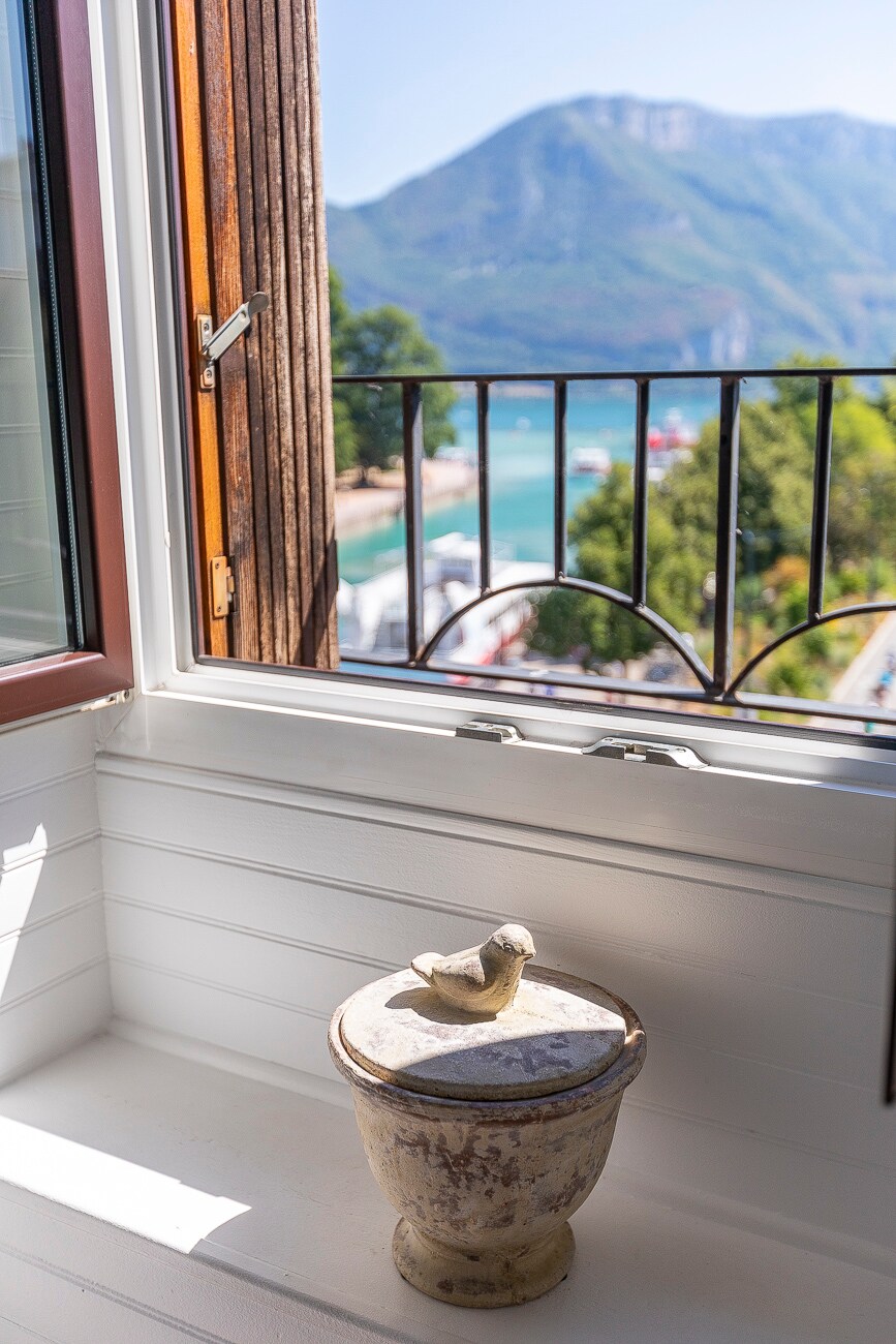 A window showcases a breathtaking view of a mountain and a glistening lake. A decorative stone container sits on the windowsill, adding a touch of character to the space. The warm light entering through the open window highlights the natural beauty outside.