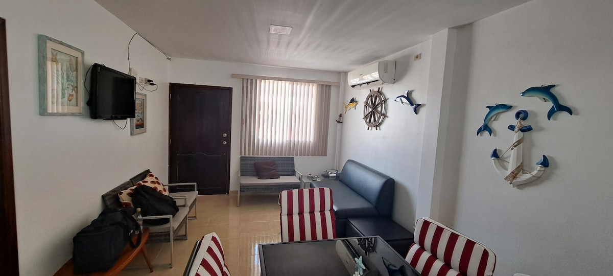 The living room features a blend of modern and nautical decor, including a dark sofa, a small seating area with striped chairs, and a wall adorned with dolphin motifs. A flat-screen television is mounted on the wall, and sunlight filters through vertical blinds.