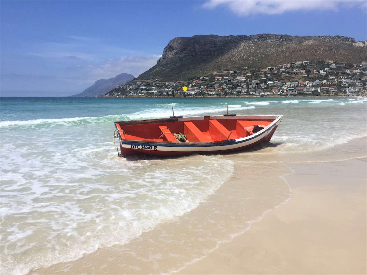 A colorful fishing boat rests in gentle waves on a sandy beach. The boat features a vibrant orange interior and a white exterior, with the backdrop of lush hillsides and residences. The water shimmers in varying shades of blue, inviting a serene seaside atmosphere.