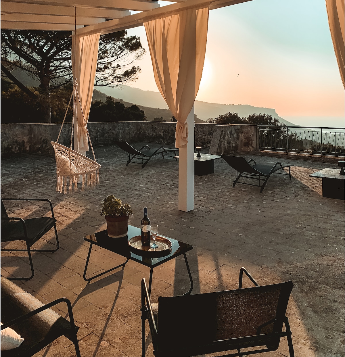 A spacious terrace is displayed, featuring comfortable lounge chairs and a small table with a bottle and glass. A swing chair is visible, while soft curtains drape elegantly. The background reveals a stunning sunset over the hills and coastline.