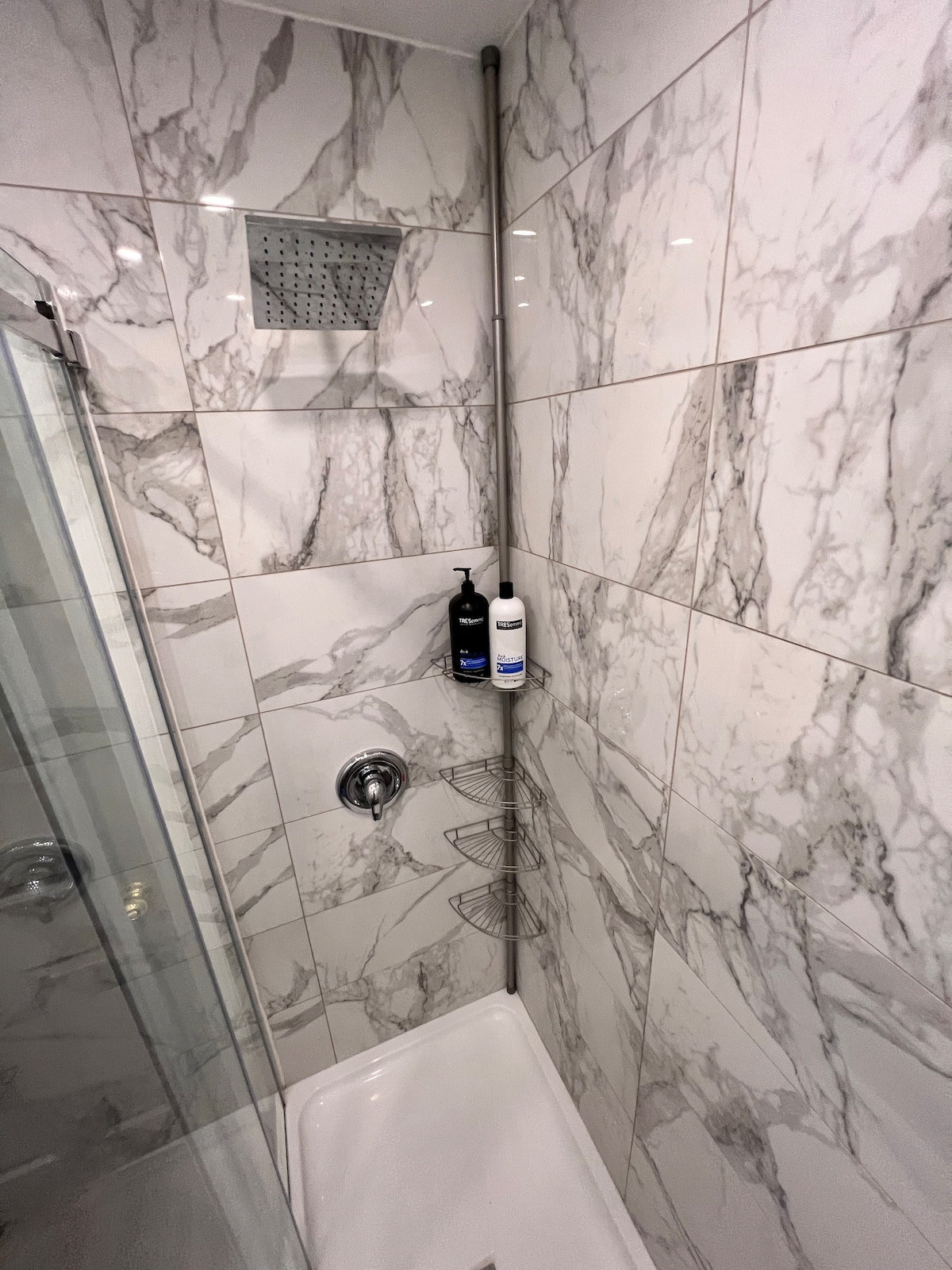 The shower area features elegant marble tiles extending from floor to ceiling. A modern showerhead is mounted overhead, complemented by wall-mounted shelves holding toiletries. A glass shower door provides an open and spacious feel.