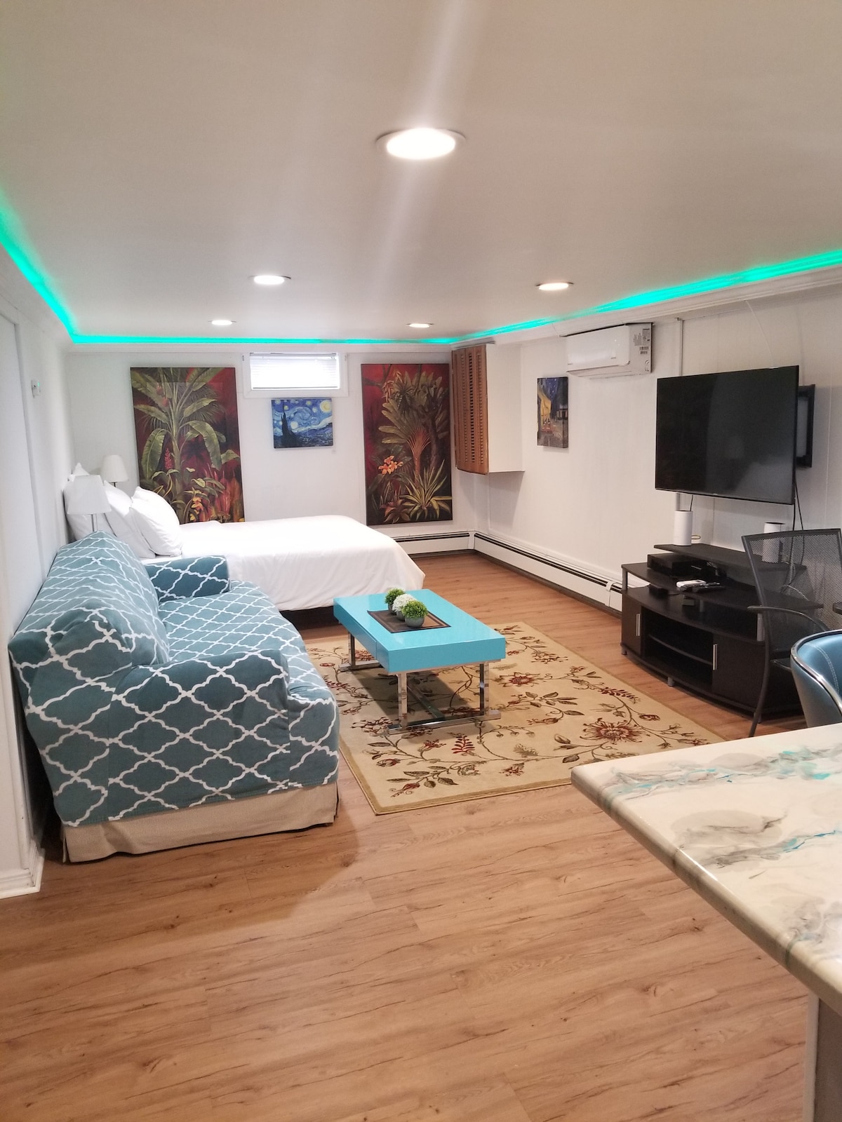 A cozy studio space features a queen-size bed with bright artwork above. A patterned couch is positioned against one wall, complemented by a coffee table. A flat-screen smart TV is mounted opposite the seating area, with LED lights lining the ceiling for subtle illumination.