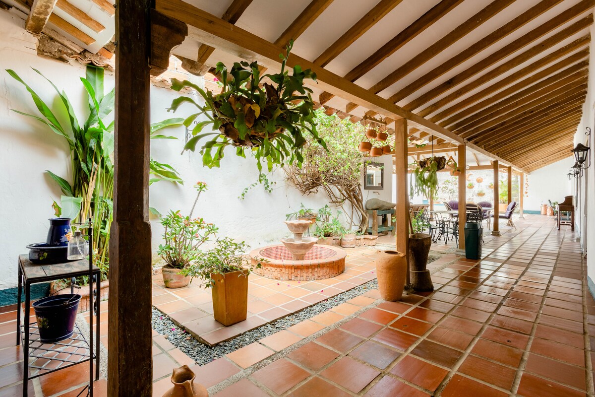 A serene corridor features a traditional clay-tiled floor and wooden beams overhead. Potted plants and decorative items line the space, leading to a central fountain surrounded by greenery. A seating area is visible in the distance, inviting relaxation in this peaceful setting.