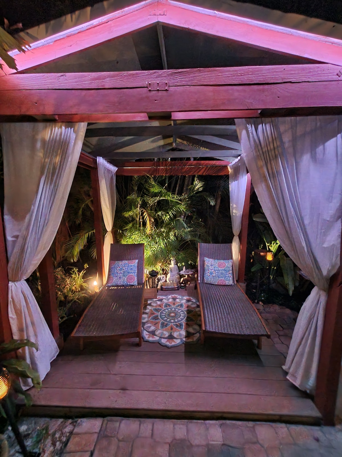 A tranquil outdoor lounging area is outlined by soft drapery, providing a serene space among lush greenery. Two wooden chaise lounges are centered on a decorative rug, with soft lighting enhancing the peaceful ambiance. A small statue adds a calming focal point.