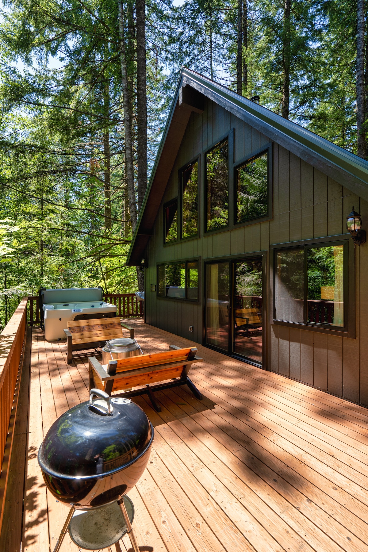 An expansive wooden deck features a hot tub and picnic-style seating, surrounded by towering trees. Large windows on the cabin offer views of the forest, while a charcoal grill is positioned near the deck edge, inviting outdoor meals in a natural setting.