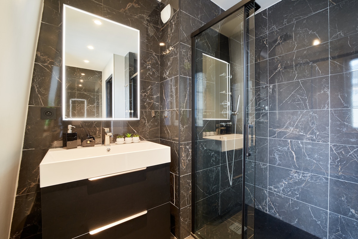 A modern bathroom features sleek black marble walls and stylish lighting. A glass shower enclosure with a handheld showerhead stands adjacent to a minimalist vanity with a sink, offering ample counter space. Essential toiletry items are neatly arranged on the countertop, complemented by decorative plants.