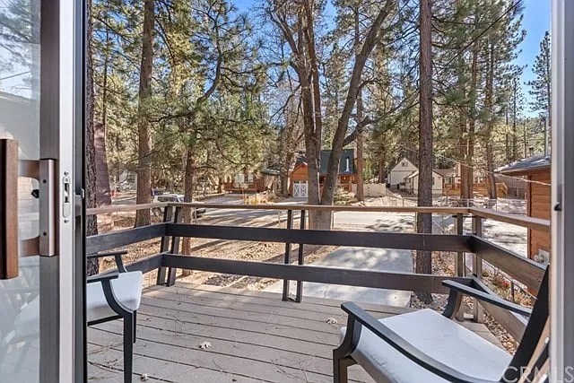 A front balcony is visible, featuring two white chairs positioned on a wooden deck. Surrounding pine trees provide a natural setting, while the view captures the tranquility of the neighborhood, showing nearby buildings and pathways below.