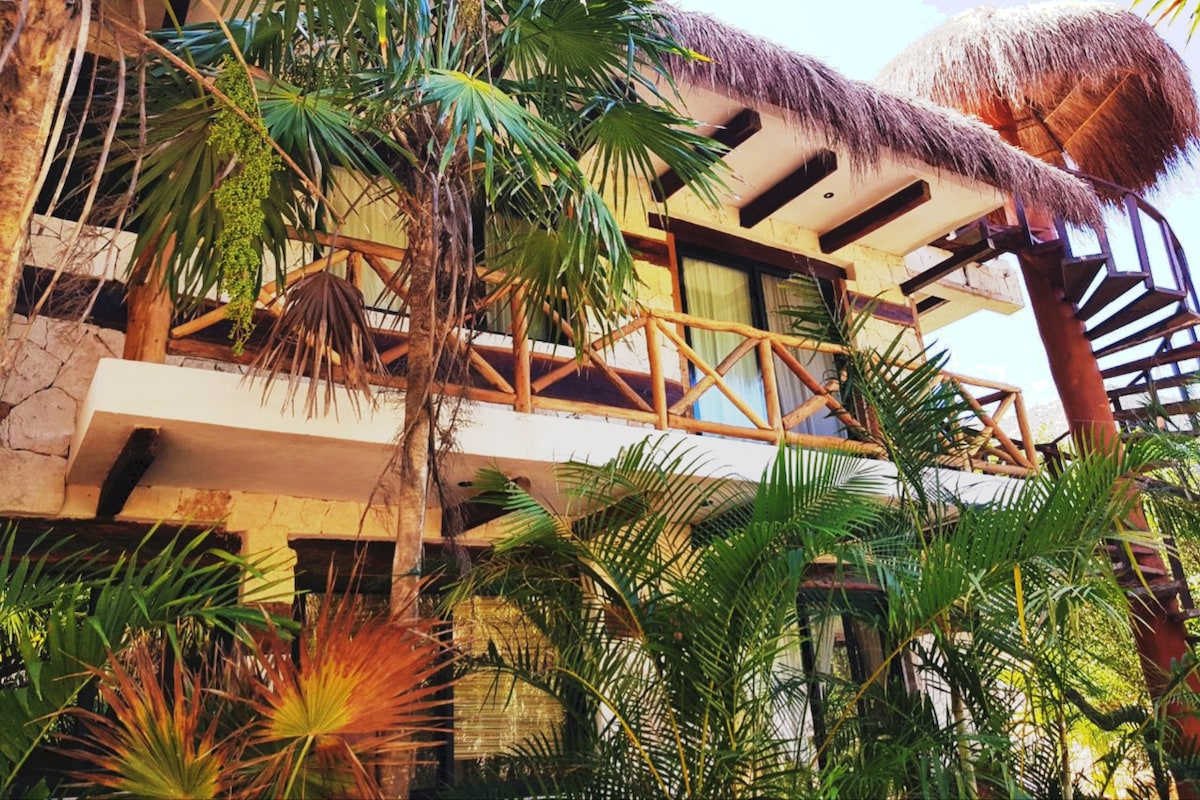 An exterior view of the villa reveals natural materials and a thatched roof, framed by lush tropical plants. A wooden balcony features a rustic railing, while a staircase leads to an upper level, enhancing the sense of the tropical setting.