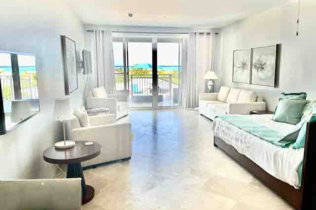 A spacious living area features large glass sliding doors that open to a balcony, allowing natural light to illuminate the room. The white furnishings include a comfortable sofa bed and armchairs, with a small side table positioned nearby. Artwork adorns the walls, and a peaceful view of the ocean is visible through the glass doors.