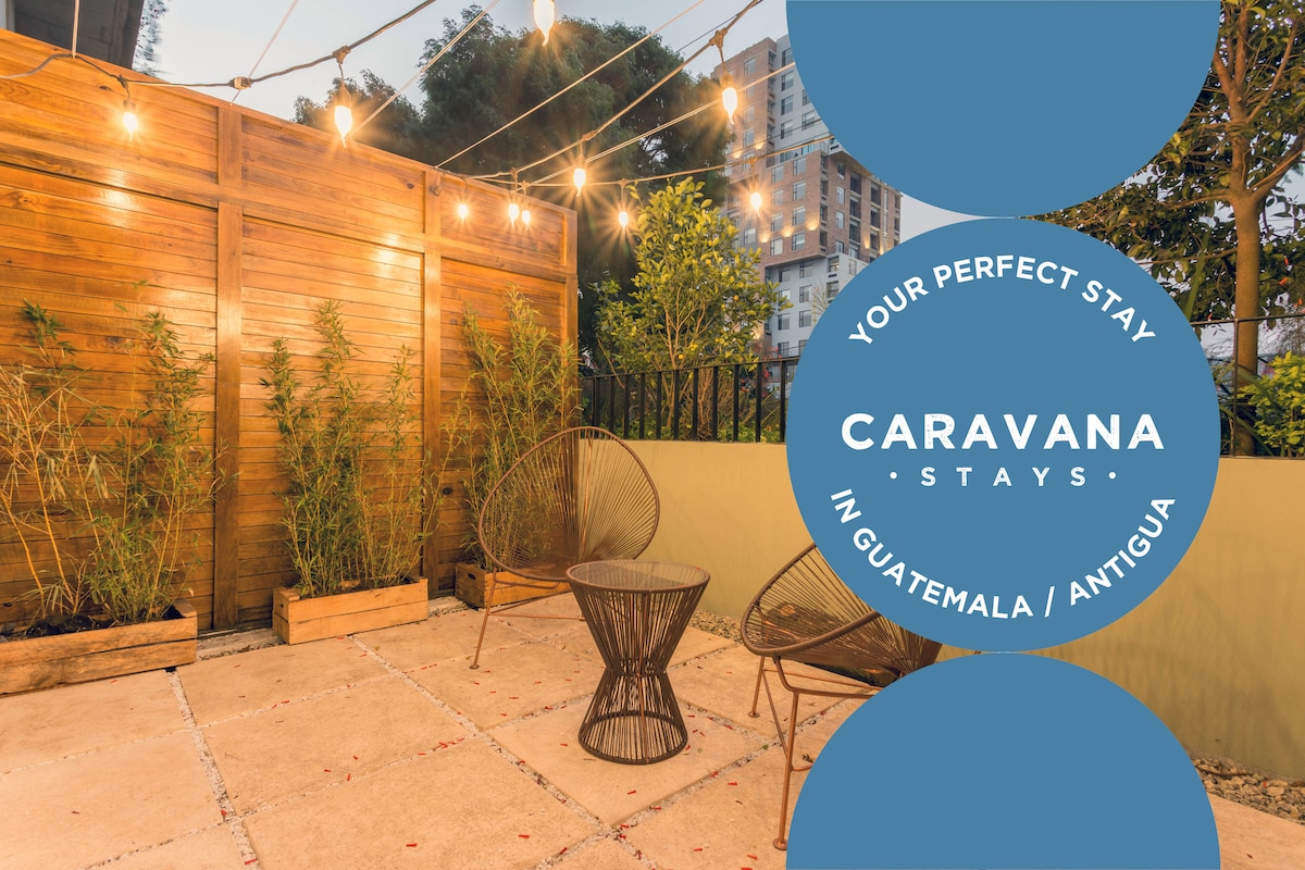 An outdoor patio features two woven chairs positioned on a tiled surface. Wooden walls are complemented by greenery in planters, and string lights hang overhead, creating a relaxed atmosphere for enjoying the space.