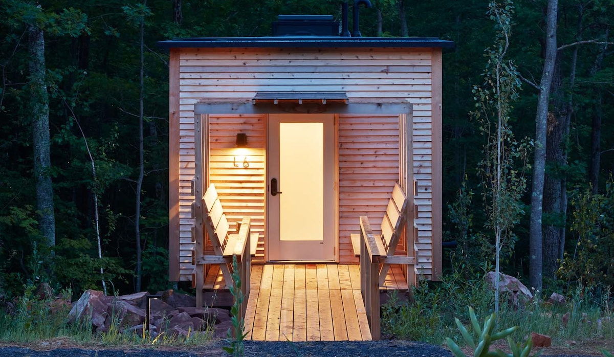 A wooden cabin is illuminated at dusk, featuring large double doors that welcome guests. A wooden deck extends towards the entrance, lined with bench seating. Surrounding trees frame the structure, enhancing the sense of seclusion and tranquility in the natural environment.