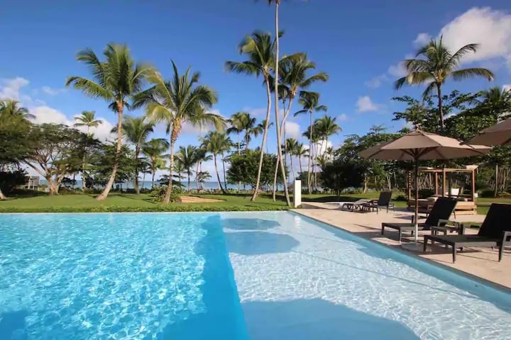 A serene pool area is shown, with clear water reflecting the blue sky. Palm trees surround the space, providing shade to the loungers situated nearby. A few umbrellas stand tall, creating comfortable spots for relaxation amidst the tranquil greenery.
