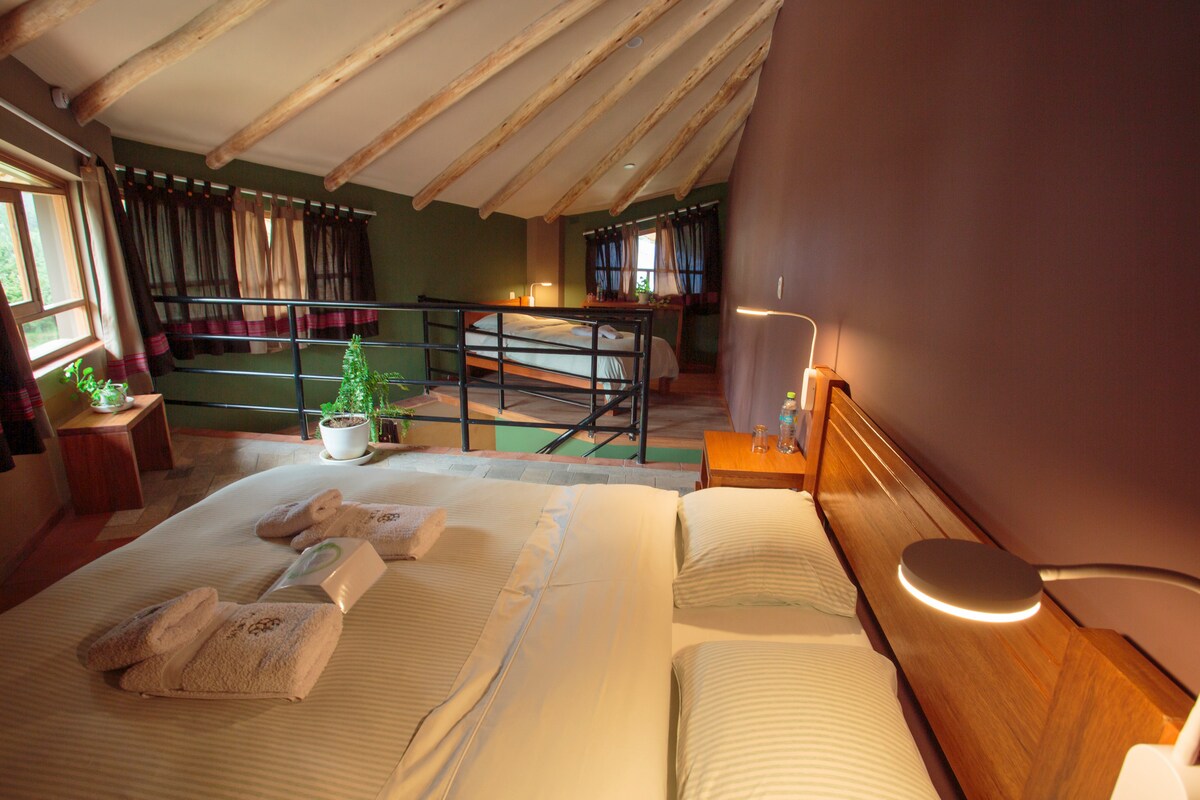 A spacious sleeping area features a double bed with light linens and decorative pillows. Soft lighting enhances the wooden accents. Large windows allow natural light to fill the room, and a small potted plant adds a touch of greenery. A cozy lounging area is visible in the background.