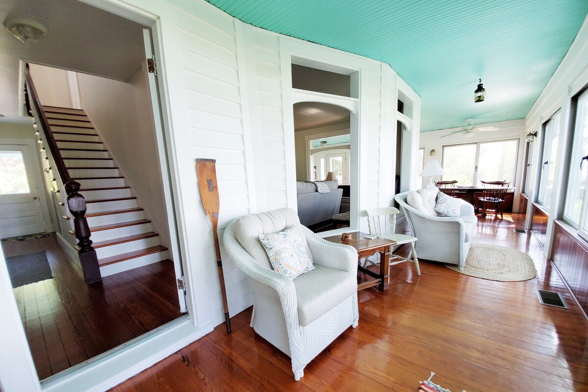 A sunlit porch features two comfortable chairs facing a small side table, providing a peaceful seating area. Light-colored walls complement the warm wooden flooring. A staircase is visible in the background, leading to additional living space.