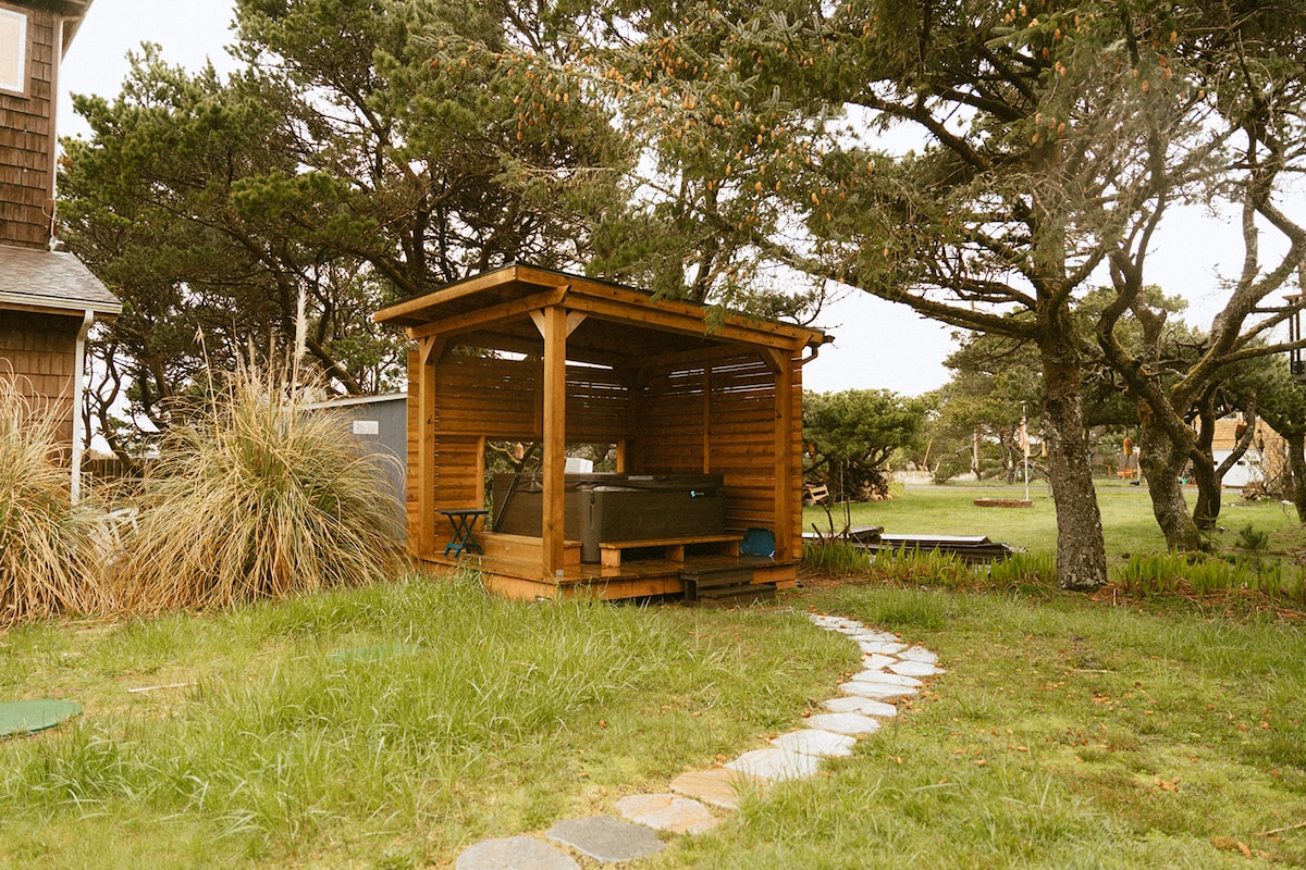 An outdoor deck area features a wooden gazebo surrounding a hot tub, with a pathway of stones leading to it. Lush grass and tall trees create a natural setting, offering a serene space for relaxation.
