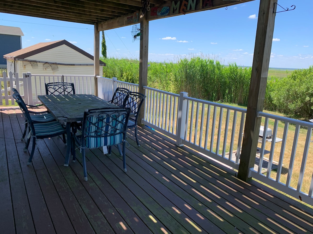 A spacious back deck features a large dining table surrounded by six chairs, overlooking lush green reeds and open bay views. Sunlight casts shadows across the wooden flooring, creating a relaxed atmosphere for family gatherings.