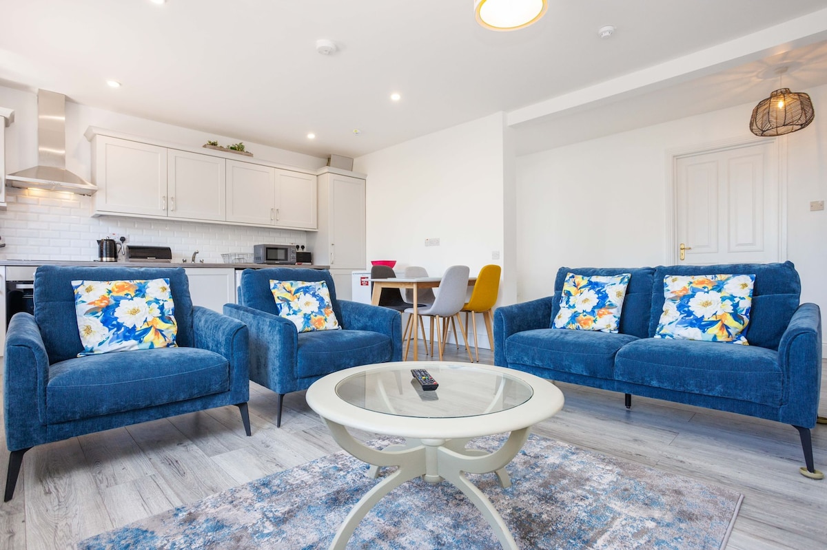 A modern living area features two blue sofas adorned with vibrant floral cushions. A round coffee table sits in the center, while a dining area with colorful chairs is visible in the background. The kitchen, with white cabinetry and stainless-steel appliances, completes the inviting space.