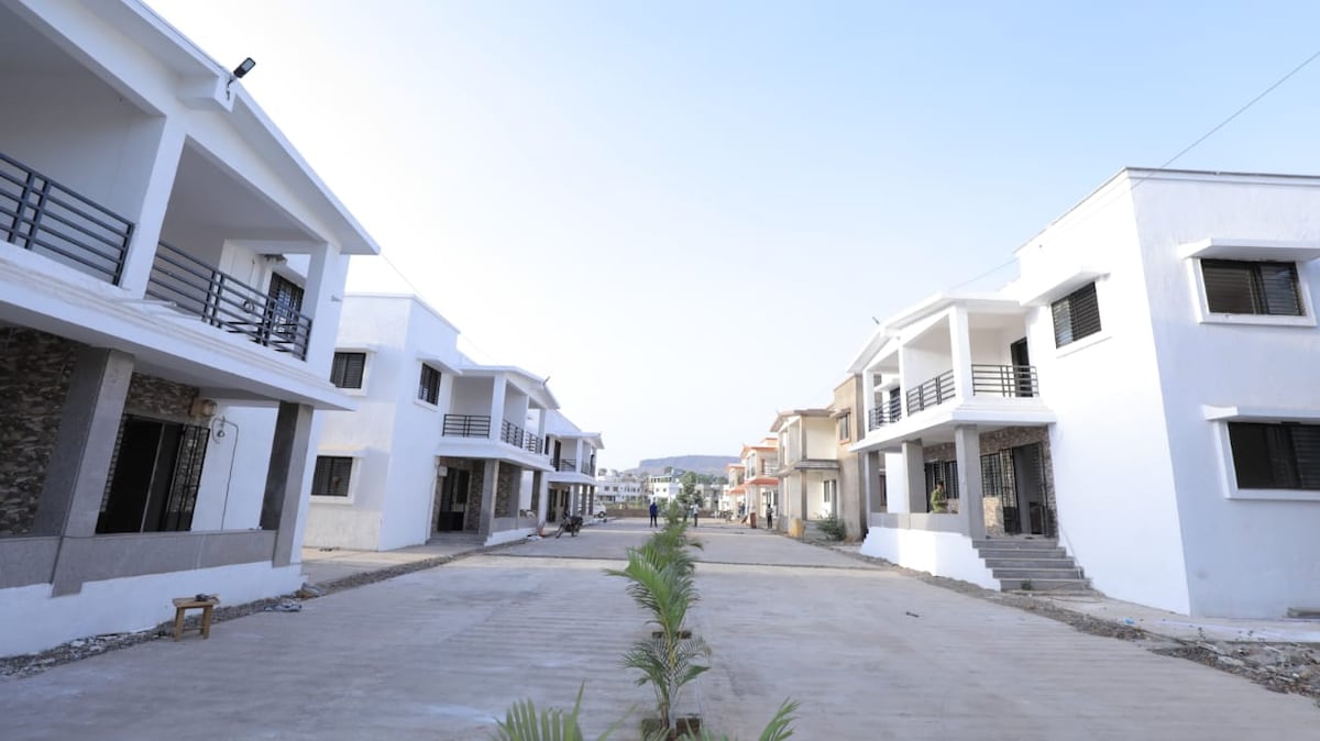 A serene street view displays two rows of modern villas, characterized by clean lines and neutral colors. The absence of vehicles suggests a peaceful atmosphere, with a landscaped area featuring small plants along the pathway, leading toward the inviting entrances of each home.