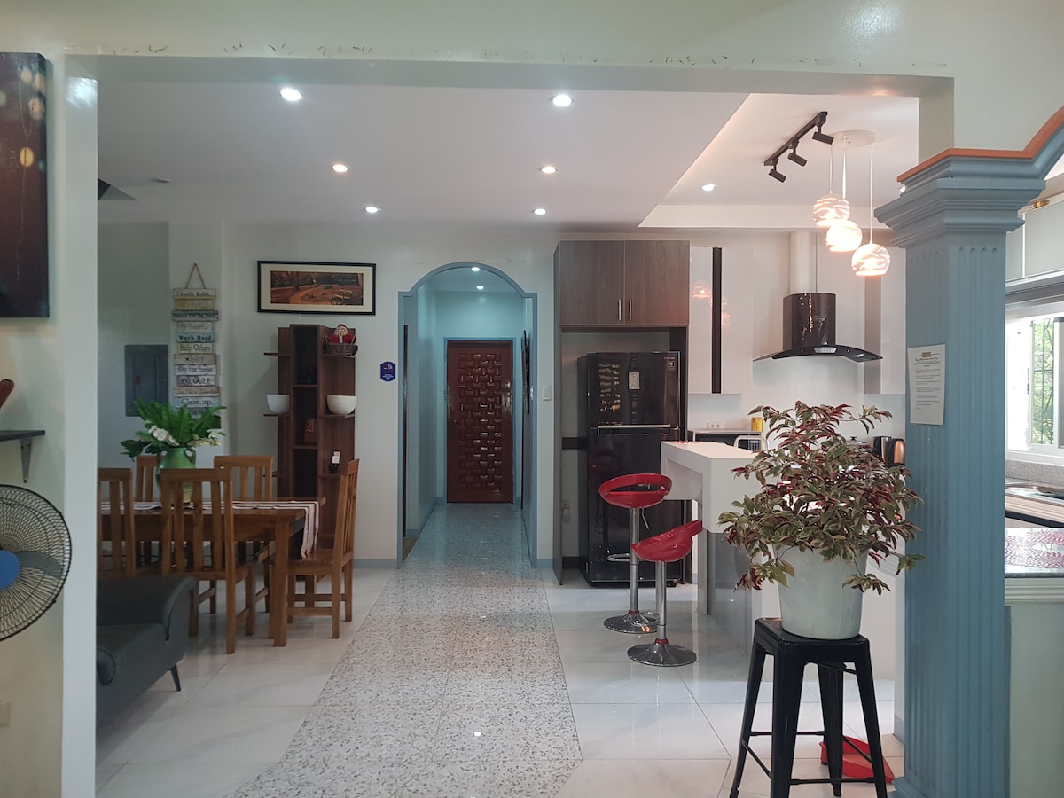 An open living and dining area is visible, featuring a wooden dining table set for six and modern bar stools at a kitchen island. Soft lighting illuminates the space, enhancing the clean lines of the cabinetry and countertops. A potted plant adds a touch of greenery.