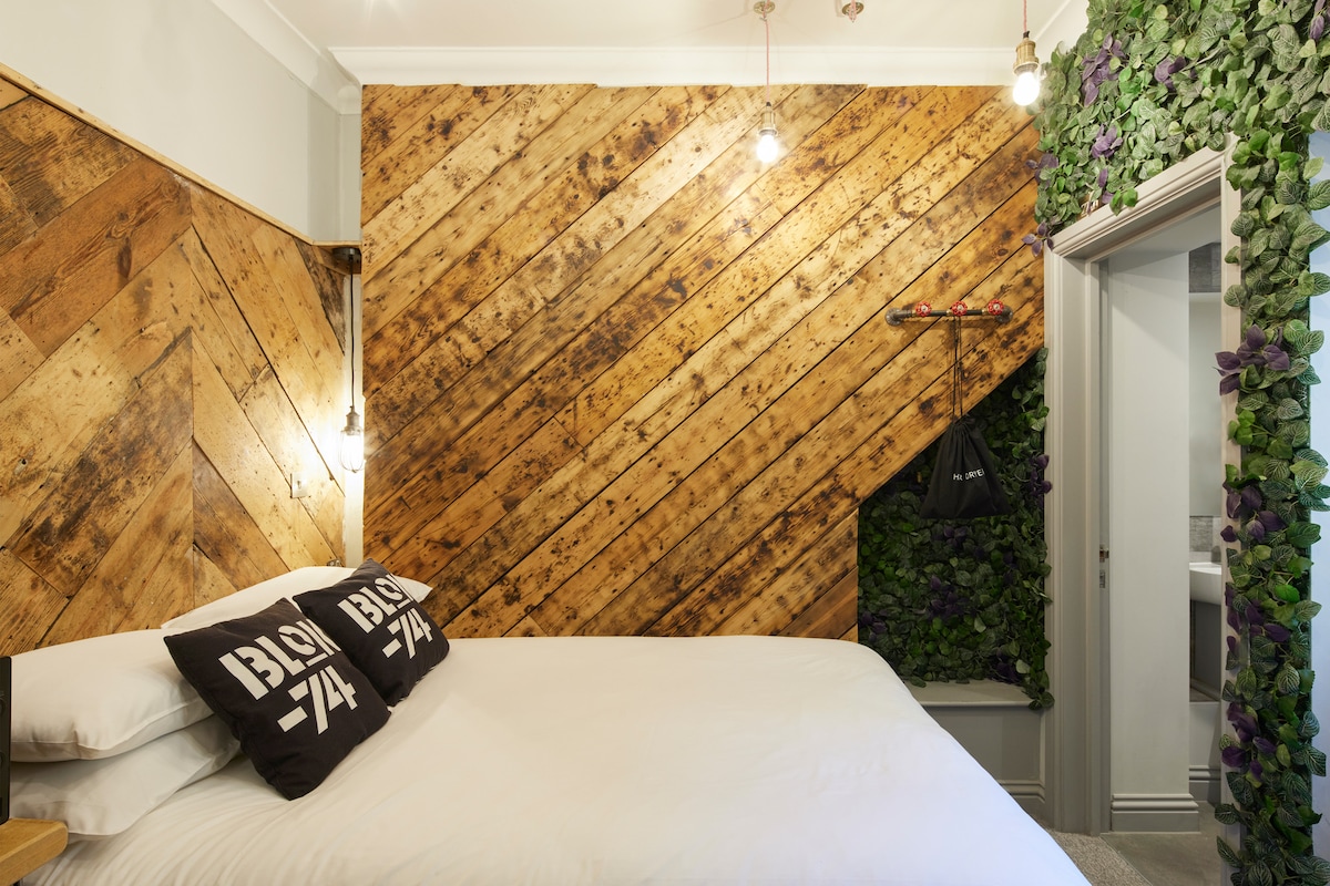 A cozy sleeping area is presented with a double bed dressed in crisp white linens and adorned with two black throw pillows. One wall features a herringbone pattern of warm wood, while another is accented by green plants, creating a refreshing atmosphere.
