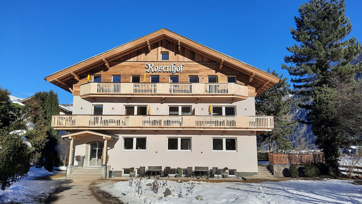 The Rosenhof building features a traditional alpine design with a wooden facade and multiple balconies. Snow covers the ground, while a clear blue sky provides a serene backdrop. The entrance is framed by landscaped greenery, creating a welcoming atmosphere.
