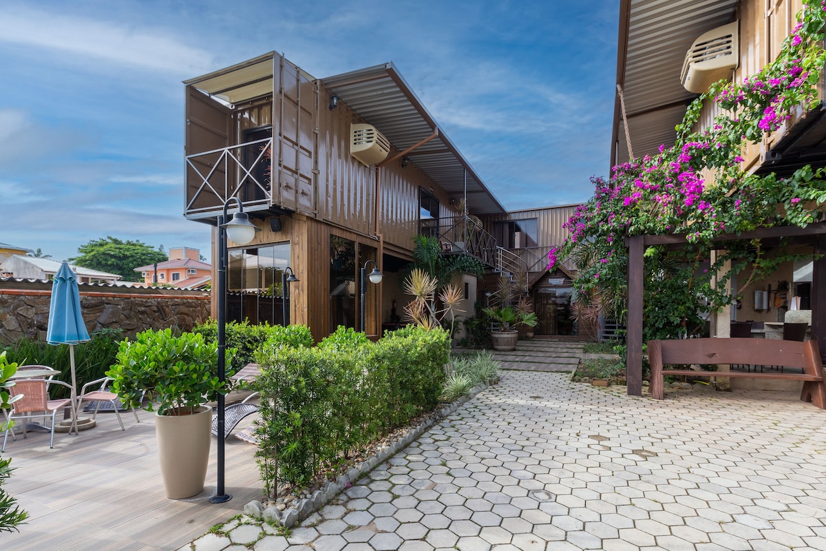 The sustainable container structure is surrounded by lush greenery and blooming flowers. Pathways are lined with patterned stones, leading to a welcoming entrance. A patio area with seating is visible, accompanied by decorative plants and a blue umbrella, creating a relaxed outdoor atmosphere.