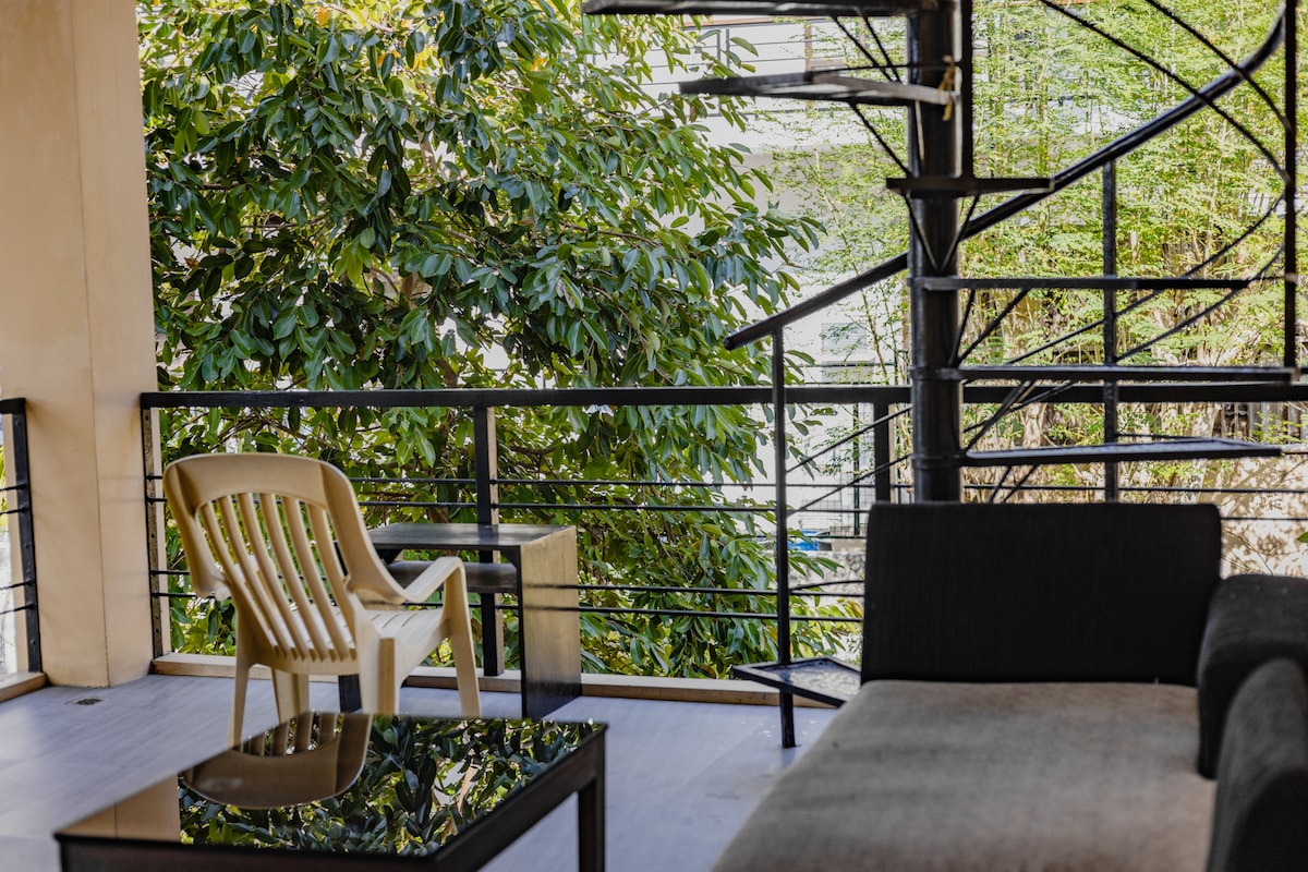 An outdoor seating area is presented, featuring a light-colored chair next to a black table, positioned under a vibrant green tree. A staircase with a black railing ascends in the background, leading to upper levels of the property.