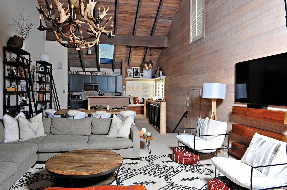 An inviting living area features a comfortable sectional sofa, a coffee table with a round top, and two accent chairs. Rustic wooden beams run across the ceiling, while a large flat-screen television is mounted on the wall, complementing the warm wooden paneling.