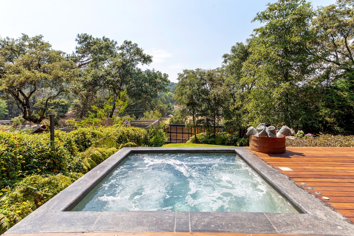 A private jacuzzi is visible amid lush greenery, providing a serene outdoor relaxation spot. Stone tiles frame the jacuzzi, while the surrounding deck offers space for seating. A tranquil view of nature and hills enhances the feeling of seclusion and peacefulness.