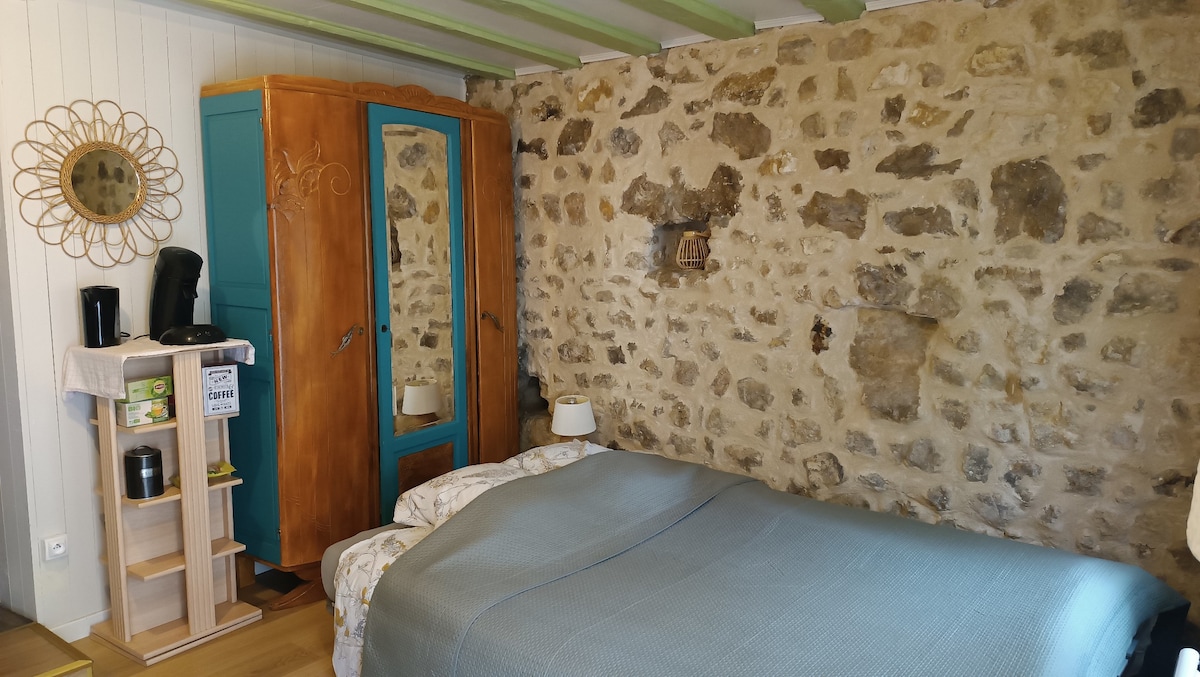 A cozy sleeping area features a spacious bed with a light blanket, complemented by stone walls that add texture. An armoire in a light wood and a vibrant blue finish stands prominently. A small table hosts a coffee maker, while a decorative mirror enhances the space.