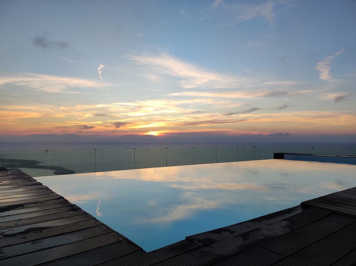 An infinity pool reflects the colorful hues of a sunset sky, creating a serene atmosphere. The pool's edge blends seamlessly with the horizon, allowing a view of the calm sea. Wooden decking surrounds the pool, inviting relaxation and leisure.