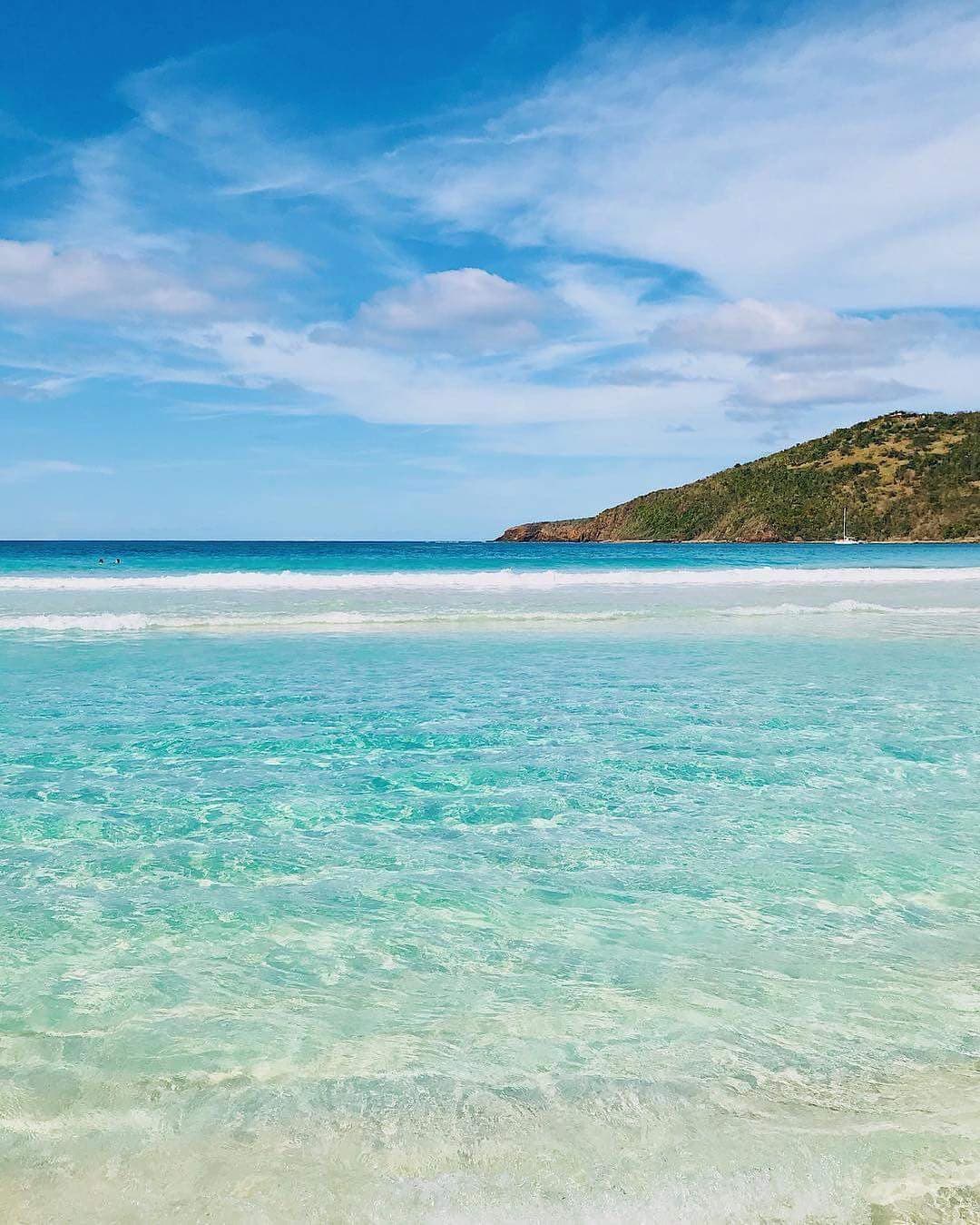 The image captures a clear, tranquil beach scene with gentle waves lapping against the shore. Shades of turquoise and aquamarine create a serene water surface, complemented by a backdrop of rolling green hills and a partly cloudy sky, inviting relaxation and natural beauty.
