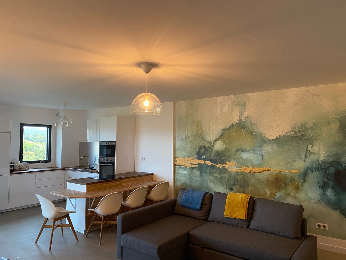 A modern living space is shown, featuring a sleek kitchen area with white cabinetry and stainless steel appliances. A wooden dining table with white chairs is illuminated by a pendant light, and a large wall is adorned with abstract artwork in calming colors.