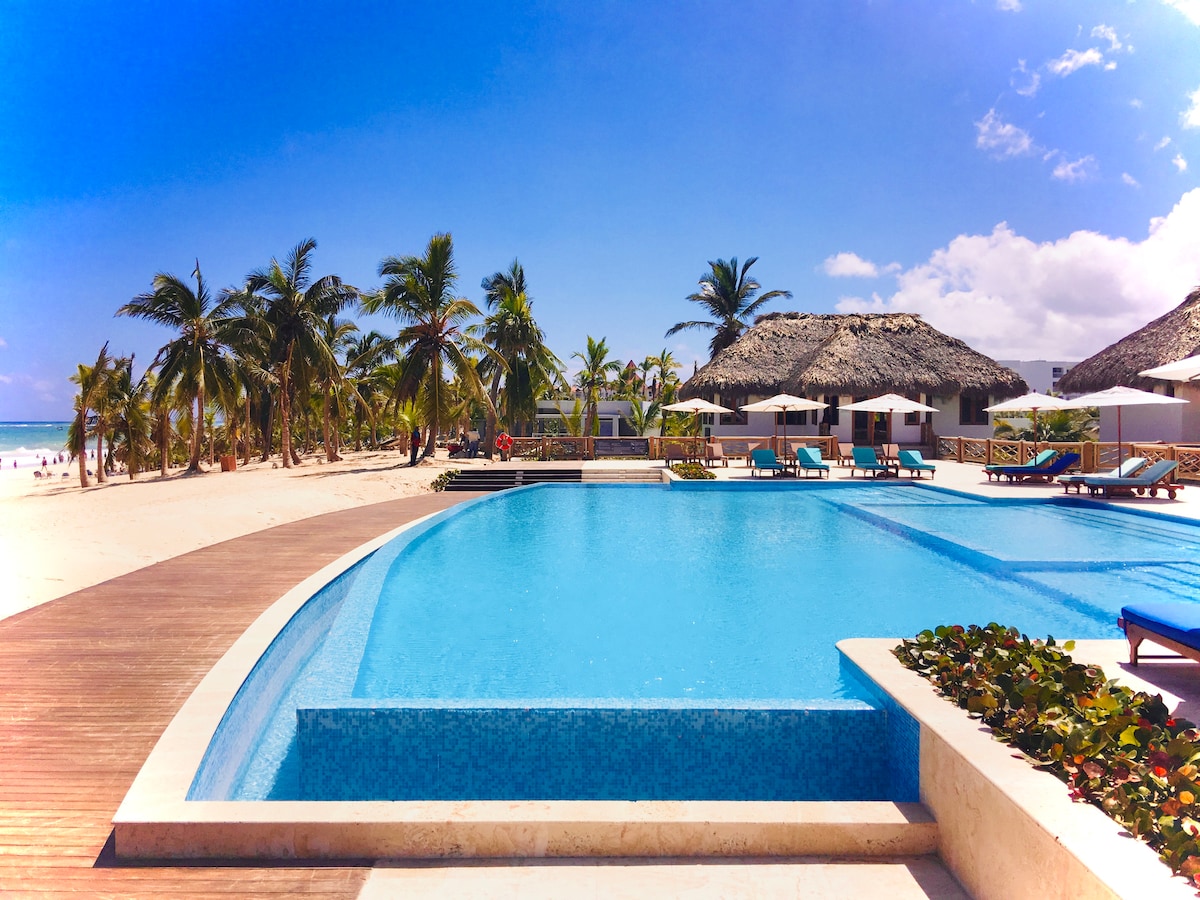 A large swimming pool is surrounded by tropical palm trees and comfortable lounge chairs. The area features a thatched-roof structure in the background, offering shade. Bright blue skies and sunlight reflect off the water, creating a serene atmosphere along the beach.