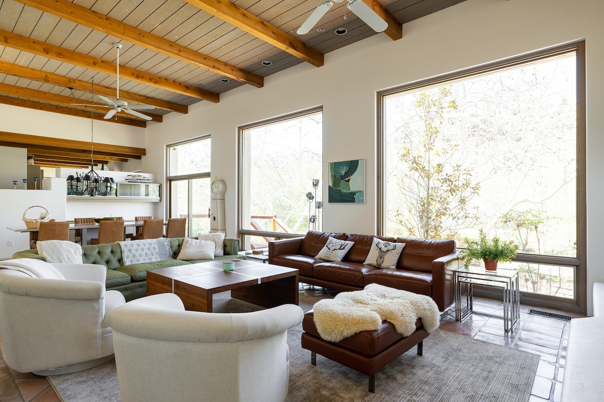 A spacious living room features high wooden ceilings with exposed beams. Large windows allow natural light to fill the room, showcasing comfortable seating arrangements including leather and fabric sofas, a wooden coffee table, and decorative plants. A soft area rug anchors the seating area.