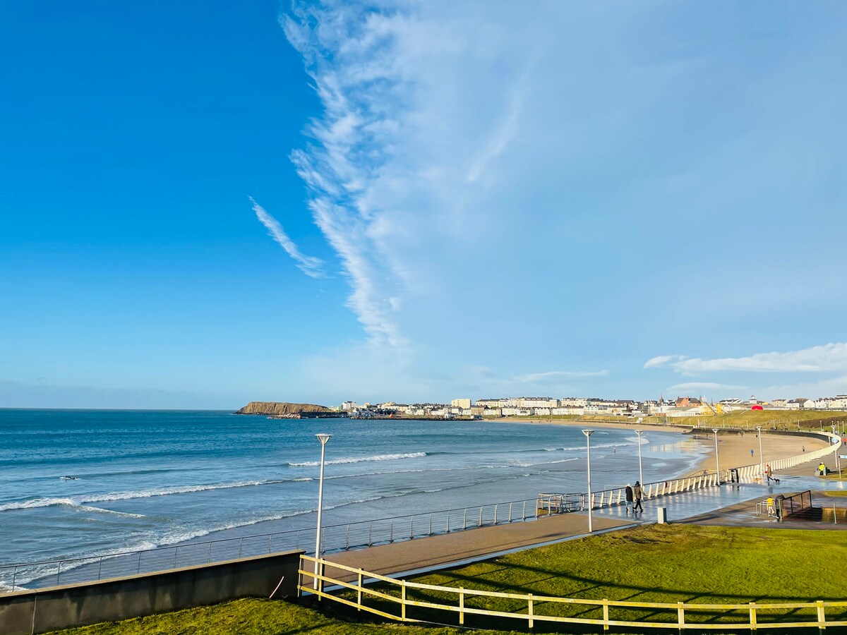 A coastal view highlights a sandy beach meeting the waves. A promenade runs alongside the shore, with gentle curves visible. Clear blue skies stretch overhead, complemented by wispy clouds. The town is visible in the distance, with varied architecture lining the coastline.