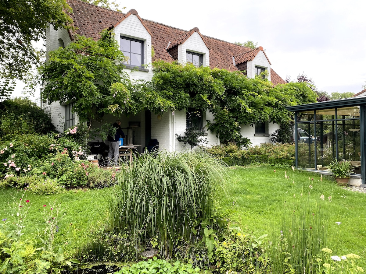 A charming cottage framed by leafy greenery is presented, featuring a covered porch with seating. The well-maintained garden includes a small pond, adding to the serene atmosphere. The home's white exterior and textured roof contribute to its inviting appearance, surrounded by flower beds and lush grass.