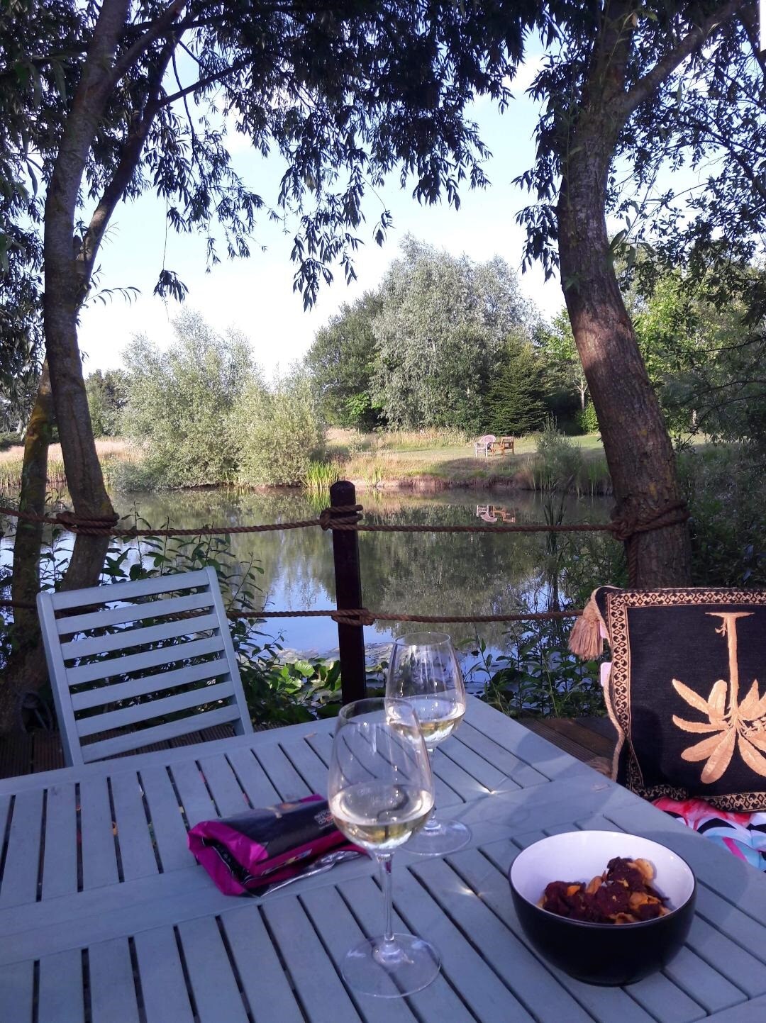 A wooden table set beside a tranquil fishing lake, with two glasses of white wine positioned on it. Lush greenery and trees frame the serene water, while a bowl of snacks sits atop the table, enhancing the inviting outdoor setting.