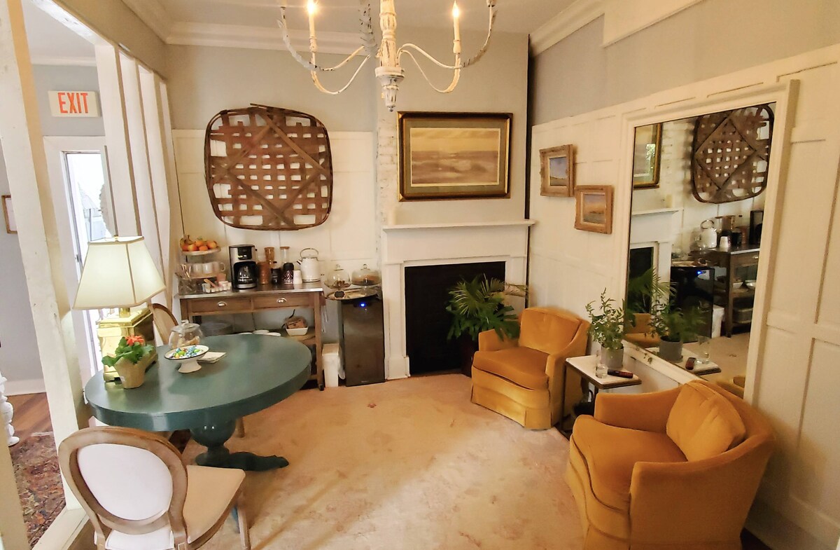 A cozy sitting area features two yellow armchairs positioned beside a round table. A fireplace is situated against a wall, framed by decorative mirrors. A cabinet holds snacks, and a plant adds a touch of greenery, contributing to the inviting ambiance.