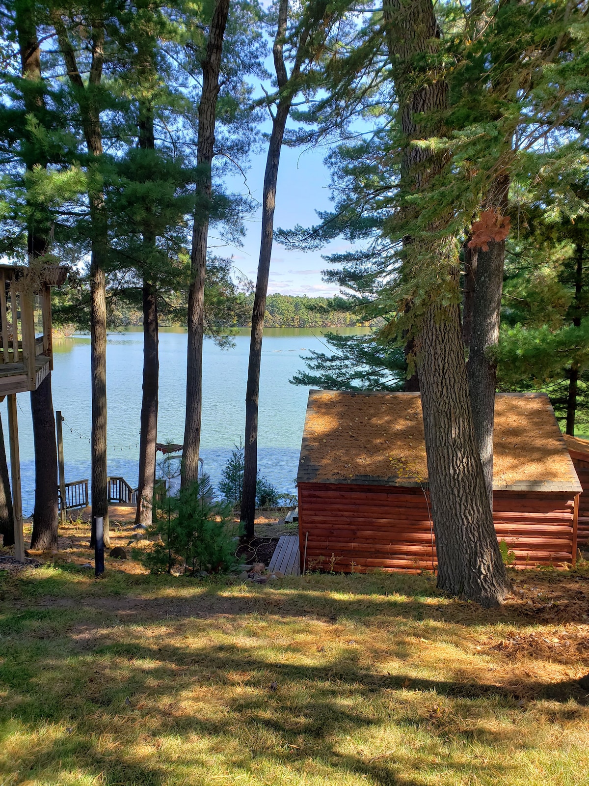 A wooden cabin is nestled among tall trees along the lakeside. The calm water reflects the blue sky, creating a serene landscape. A wooden dock is visible nearby, adding to the natural setting. The area is surrounded by lush greenery, enhancing the peaceful ambiance.