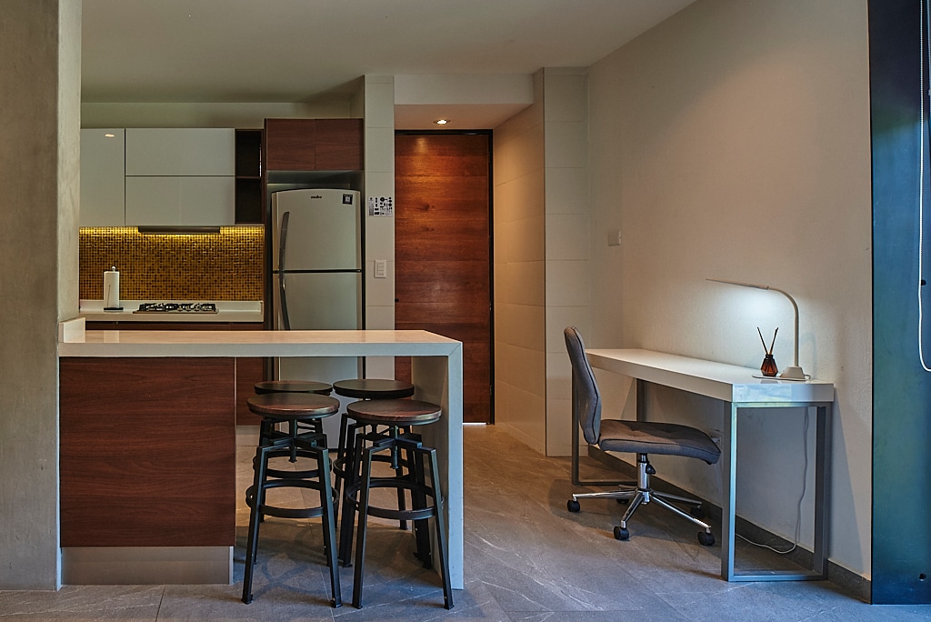 A modern kitchen area features sleek cabinetry and stainless steel appliances. A breakfast bar with three stools is positioned beside a small workspace that includes a desk and an office chair. Soft lighting enhances the clean lines of the design.