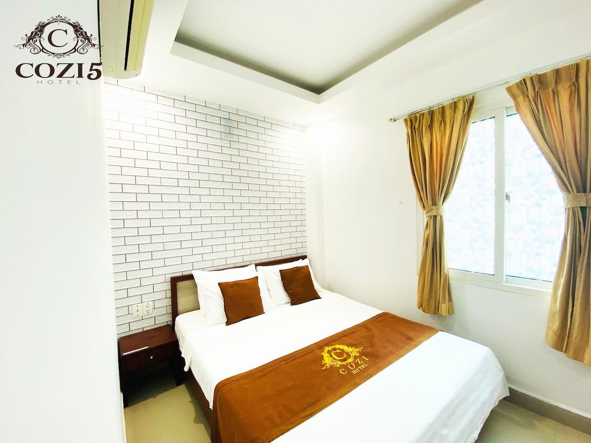A bright sleeping area is presented with a spacious bed dressed in white linens and a warm-toned blanket. Two accent pillows are positioned at the head of the bed. Natural light filters in through a large window, which is framed by soft curtains.