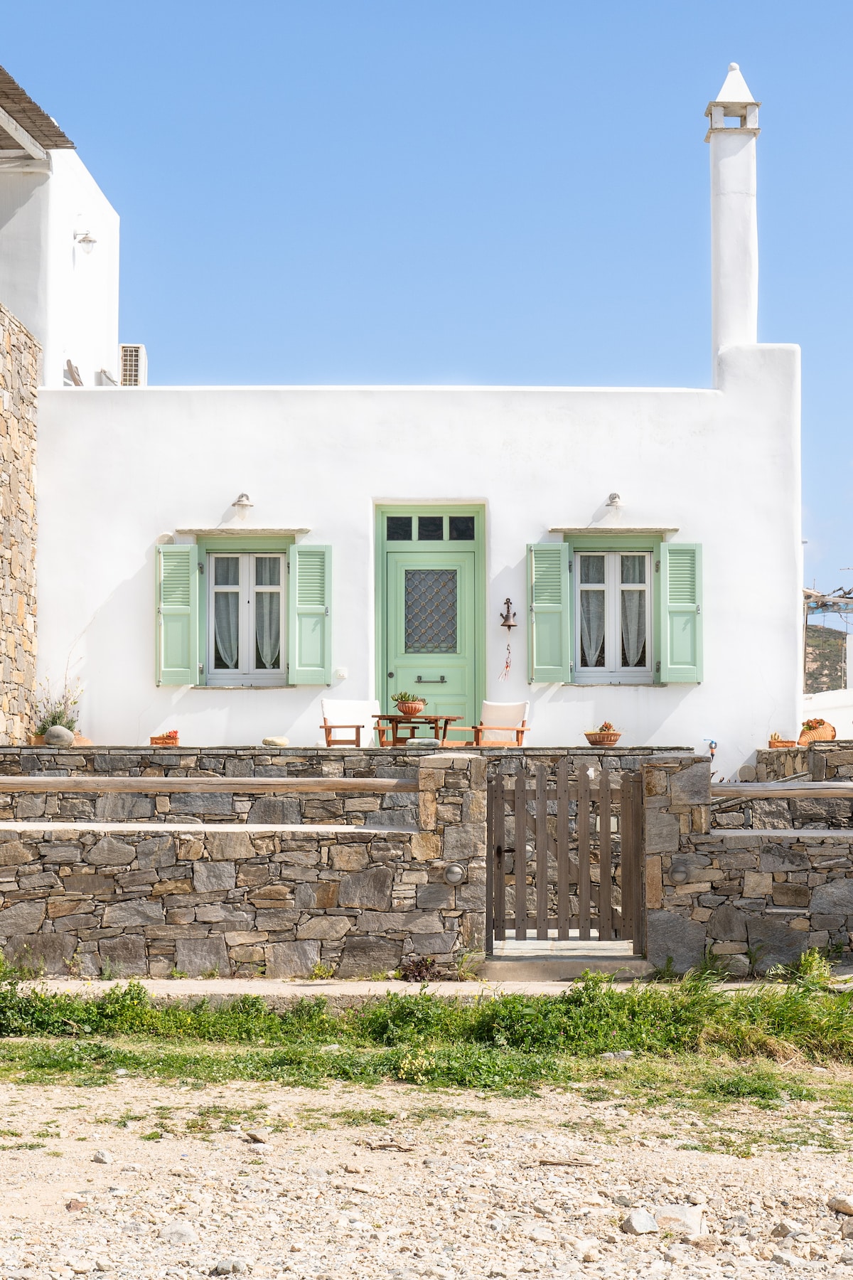 A charming white building features green shutters and a front door with decorative elements. A small outdoor seating area is visible, with a table and chairs placed on the porch. Stone walls and a wooden gate frame the entrance, providing a welcoming view.