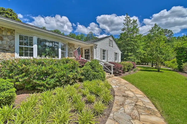 A welcoming entrance to a single-story house is framed by a well-maintained stone walkway and vibrant green landscaping. Windows adorned with white shutters reflect natural light. The garden features varied shrubs and trees, contributing to the tranquil setting.