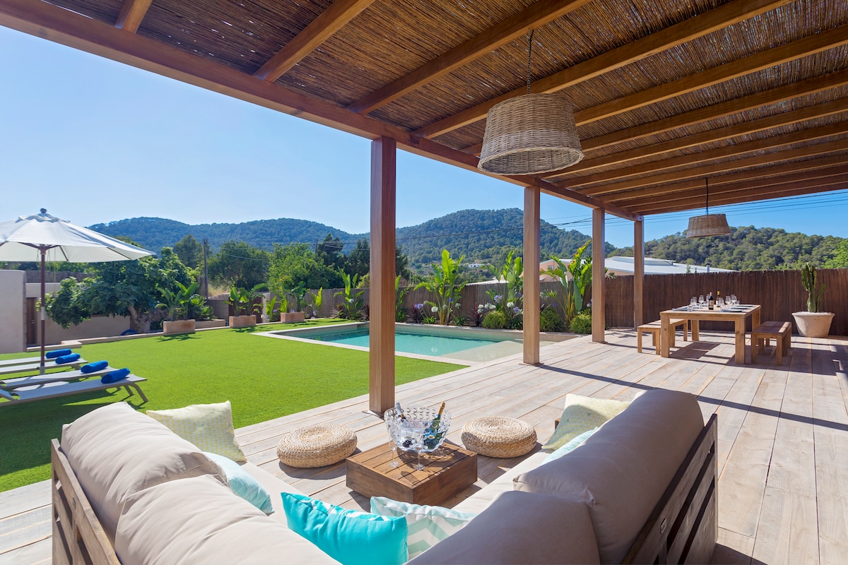 An outdoor seating area features a large sofa with light cushions, offering a view of the green lawn and a sparkling pool. A dining table stands nearby under a wooden pergola, with sun loungers arranged beside the pool, surrounded by lush plants and hills in the distance.