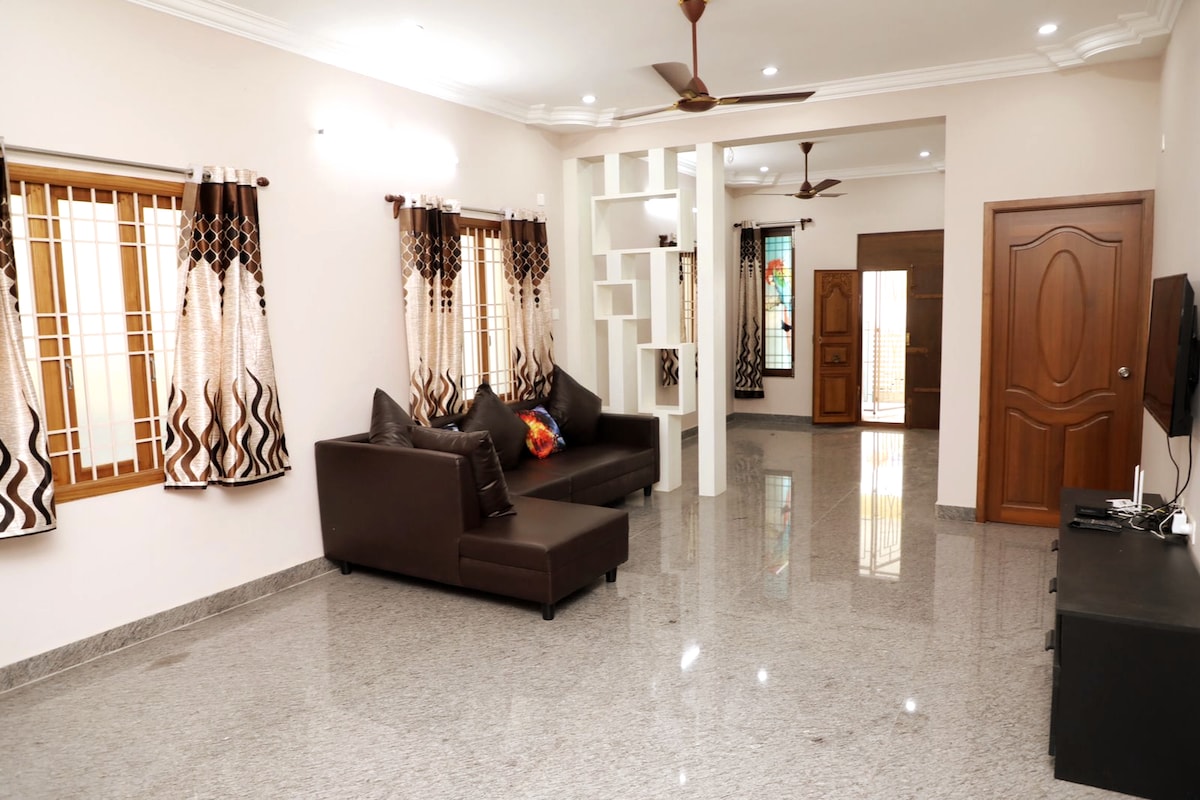 A spacious living area features polished granite flooring and large windows with light-filtering curtains. Comfortable seating is provided by a dark sectional sofa. Decorative shelving units are situated against the wall, while a door leads to additional rooms, enhancing the open feel of the space.