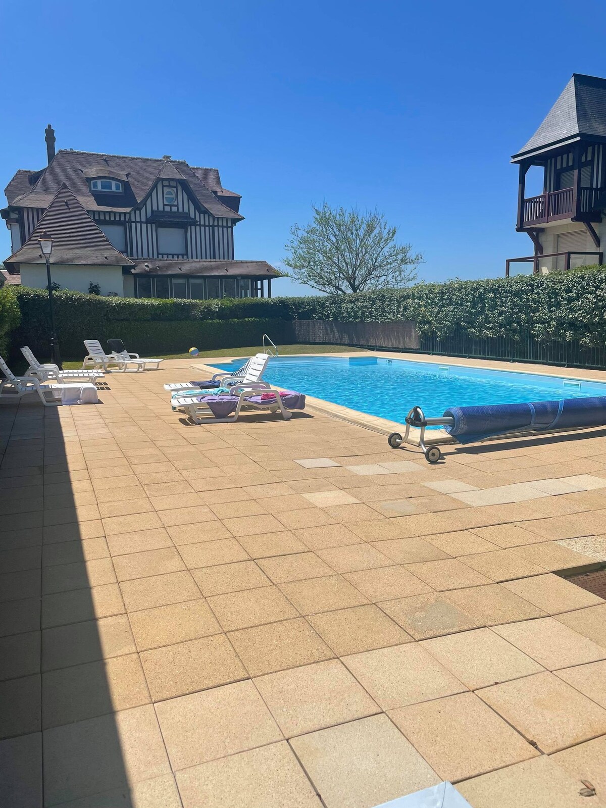 A serene outdoor area features a swimming pool with clear blue water, surrounded by tiled decking. Several lounge chairs are positioned around the pool. Lush greenery provides privacy, and two distinctive houses are visible in the background under a clear blue sky.