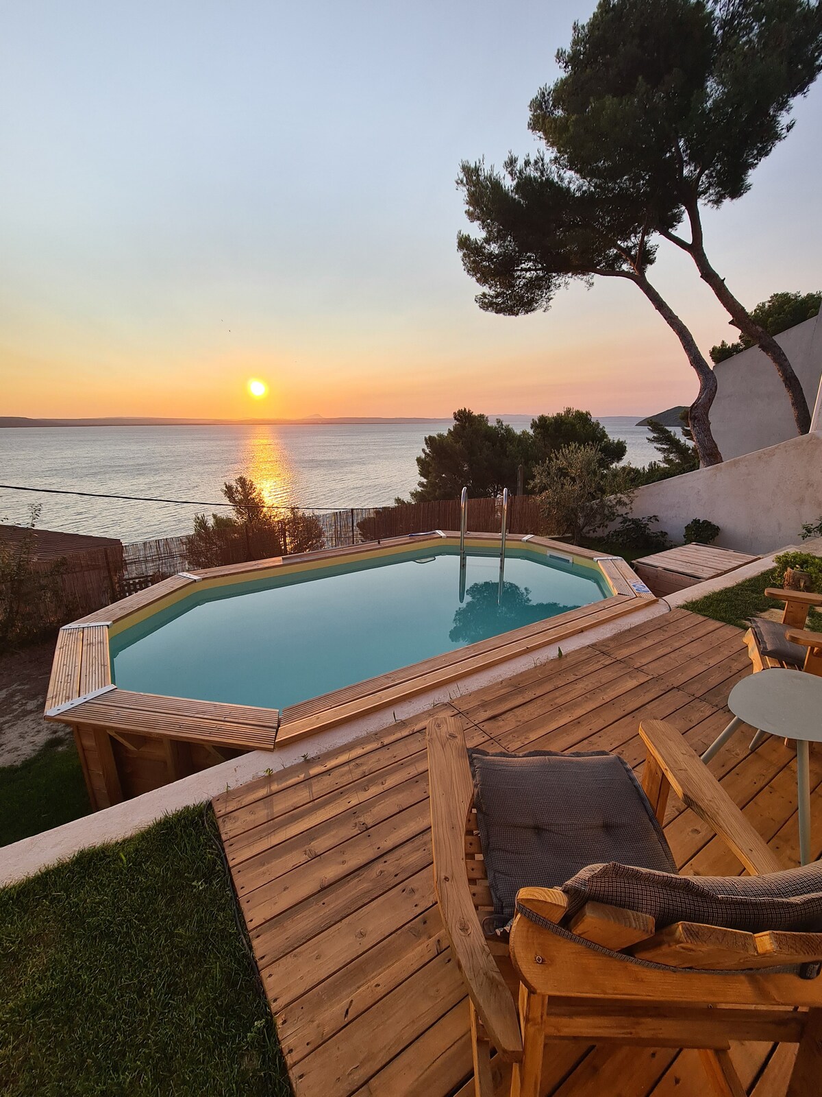 A serene view of the sunset over the water is captured from a wooden deck featuring a small pool. Two wooden chairs and a round table sit comfortably on the deck, surrounded by lush greenery and tranquil surroundings.