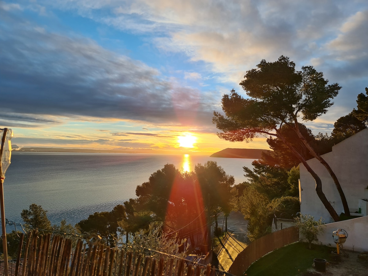 A serene sunset is captured over the water, with hues of gold and orange reflecting on the calm surface. Silhouettes of pine trees are visible against the colorful sky, offering a peaceful backdrop to the scenic view. The landscape invites relaxation and tranquility.